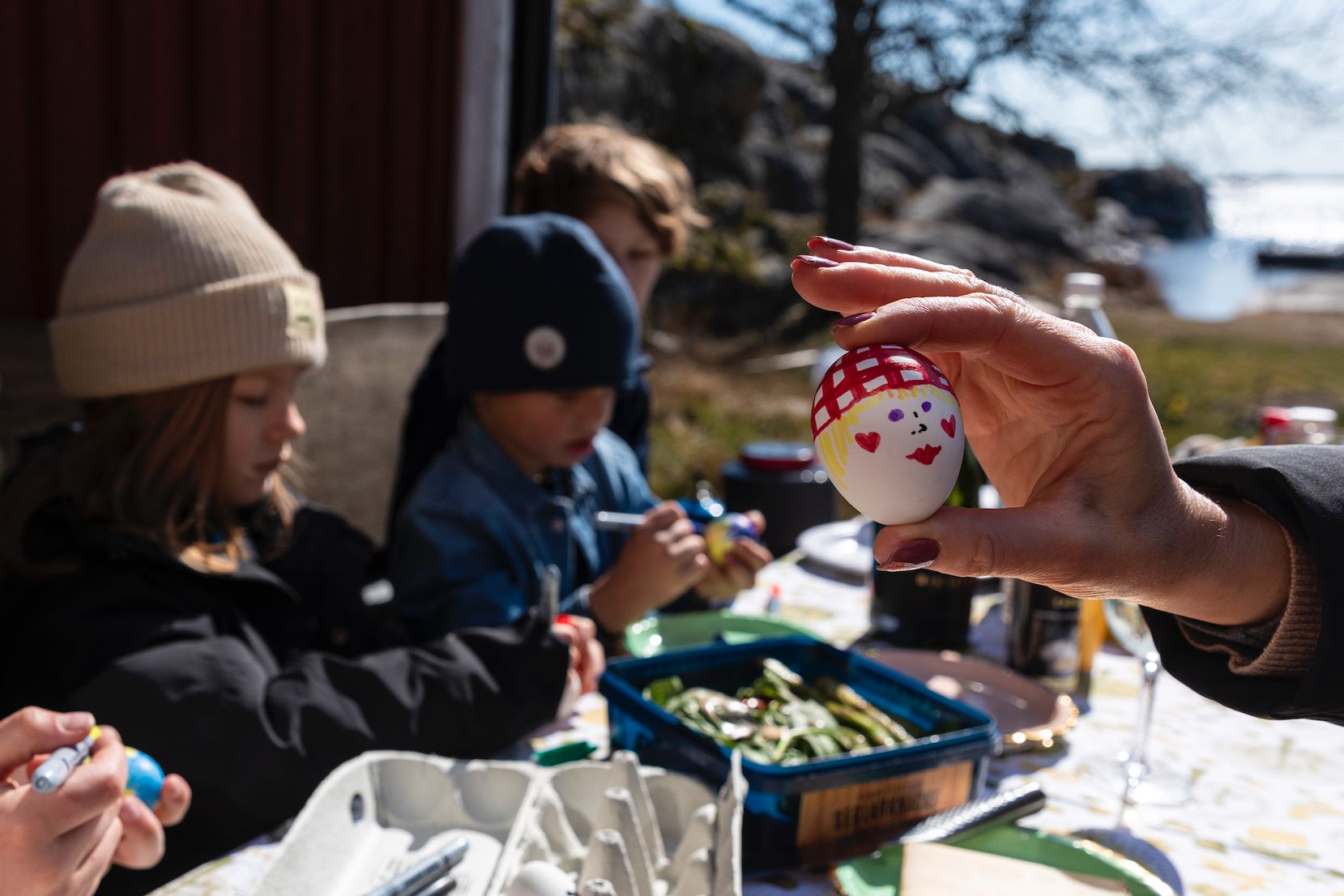 En hand håller ett målat påskägg framför barn som målar ägg.