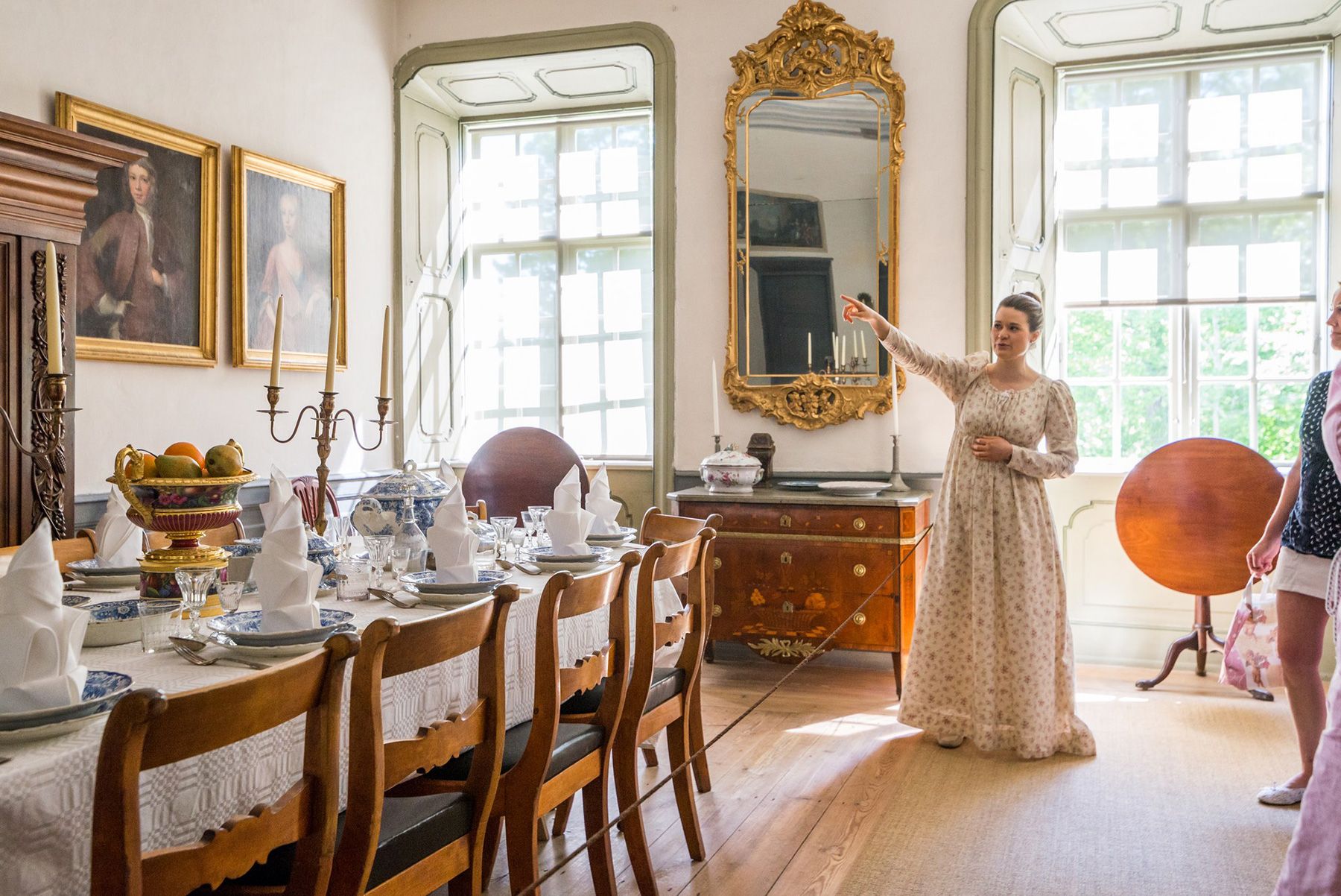 A tour guide dressed in a long white noblewoman's dress at Louhisaari Manor shares stories about the history of the dining room. There are paintings and mirrors on the walls and the table is laid beautifully.