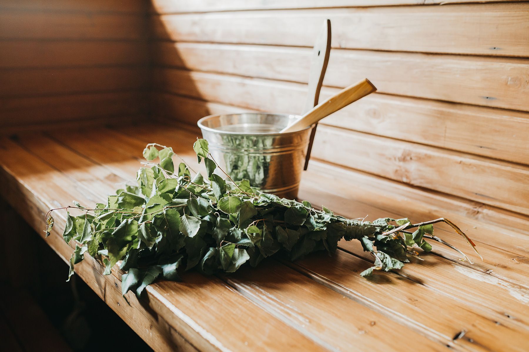 A bucket, ladle, and birch leaves on a bench inside a sauna.