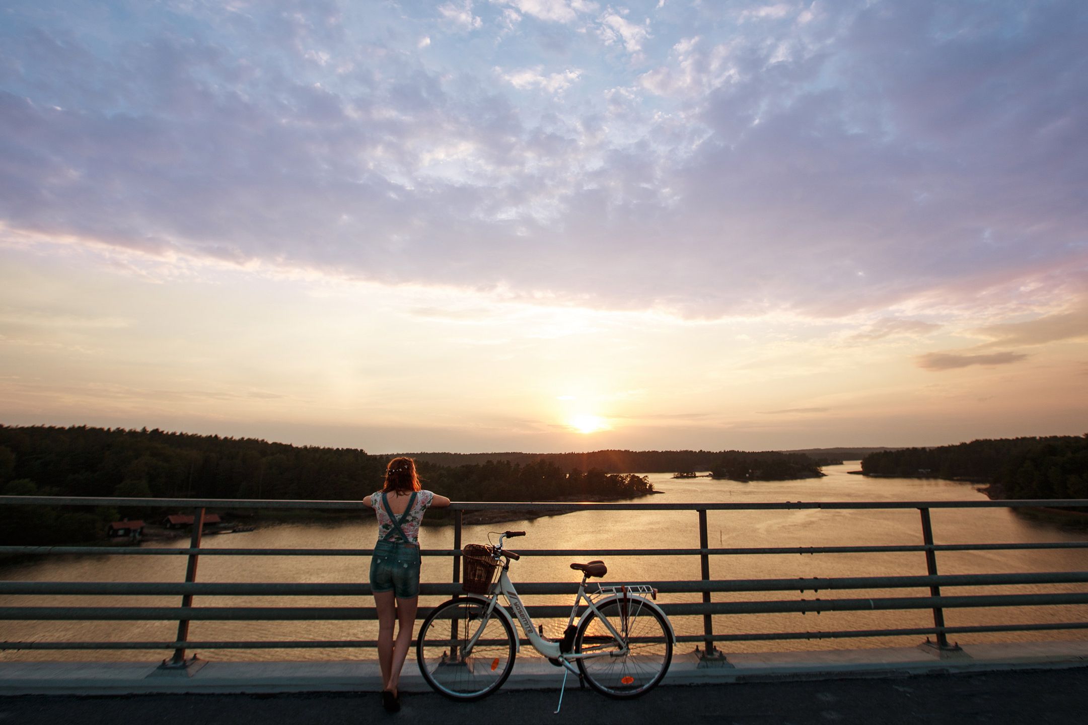 Nainen seisoo sillalla polkupyöränsä vieressä ja nojaa kaiteeseen. Hän katsoo kohti saaristoa ja auringonlaskua, joka on värjännyt meren oranssiksi.
