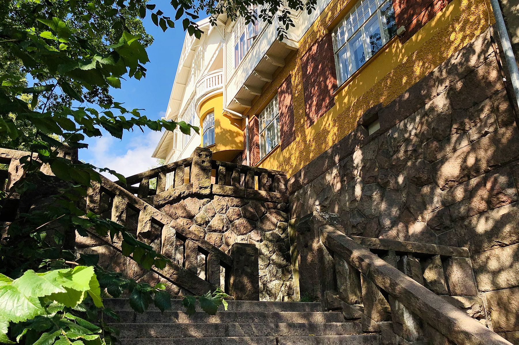 The meandering Muistainveljestenkuja Way stairs in the shade of the trees. At the top of the stairs is a yellow Art Nouveau building.
