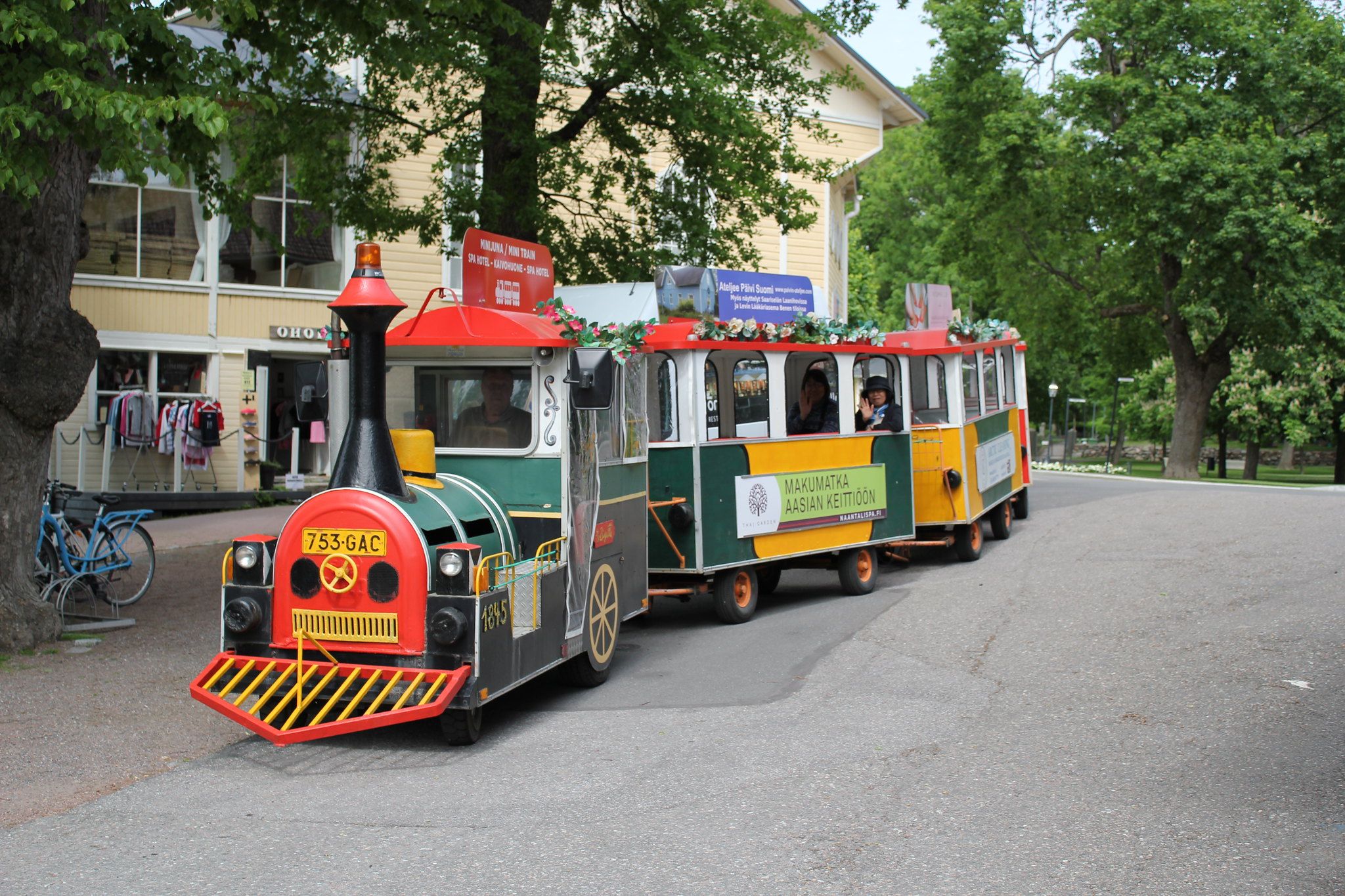 A colourful miniature train travels through the centre of Naantali. There are a few passengers along for the ride and there is a green park in the background.